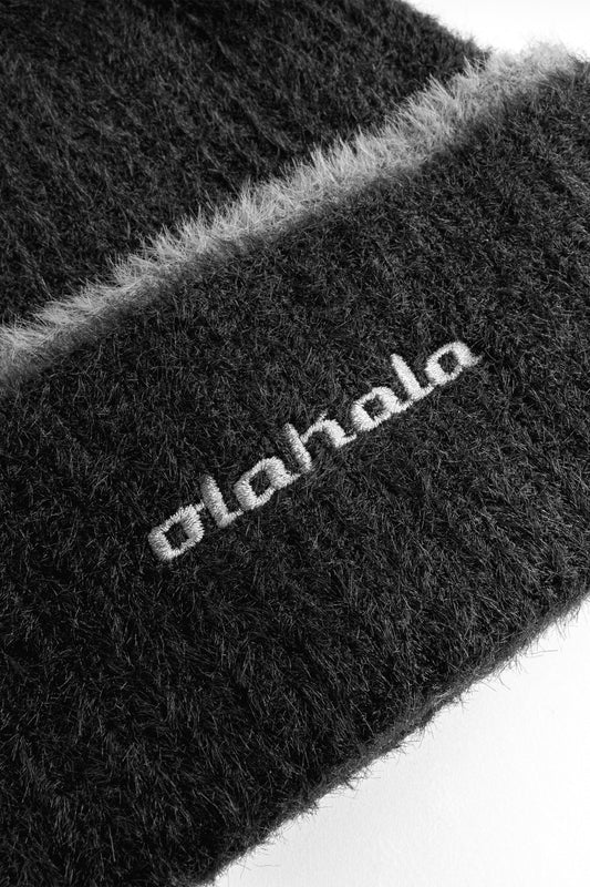 Nahaufnahme einer schwarzen und weißen Teppichstruktur, die den Hintergrund für die Fuzzy Strick-Beanie mit umgeschlagenem Rand und gesticktem OLAKALA Logo bildet. Unisex Design.