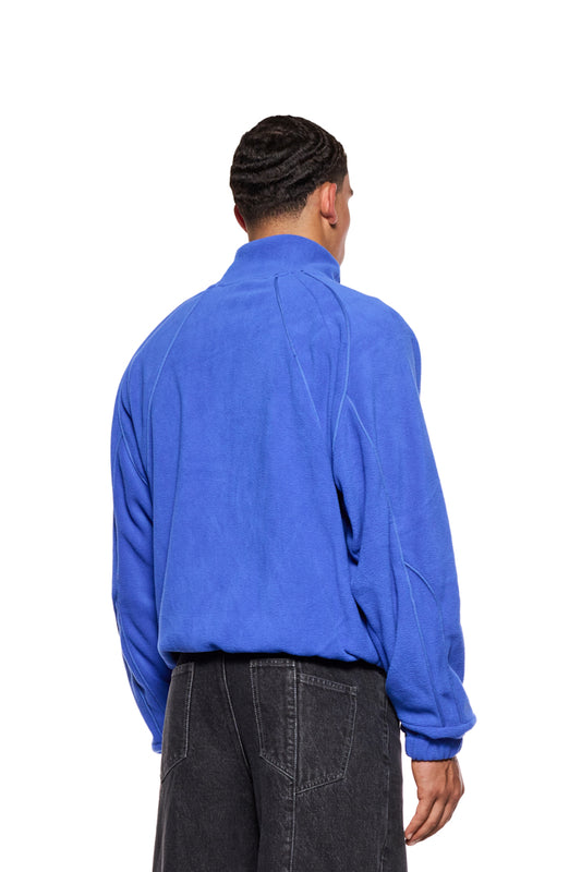 Mann trägt Fleece Jacket Dazzling Blue mit Teilungsnähten, gesticktem OLAKALA-Logo und Eingriffstaschen, Oversized-Passform aus weichem Fleece-Stoff.