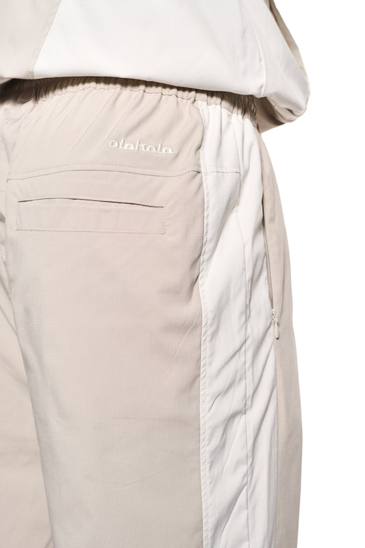 Person trägt OLAKALA Sports Tracksuit Pants Sand mit Teilungsnähten, Reißverschlusstaschen und gesticktem Logo, aus robustem Rip-Stop-Gewebe, lockere Passform.
