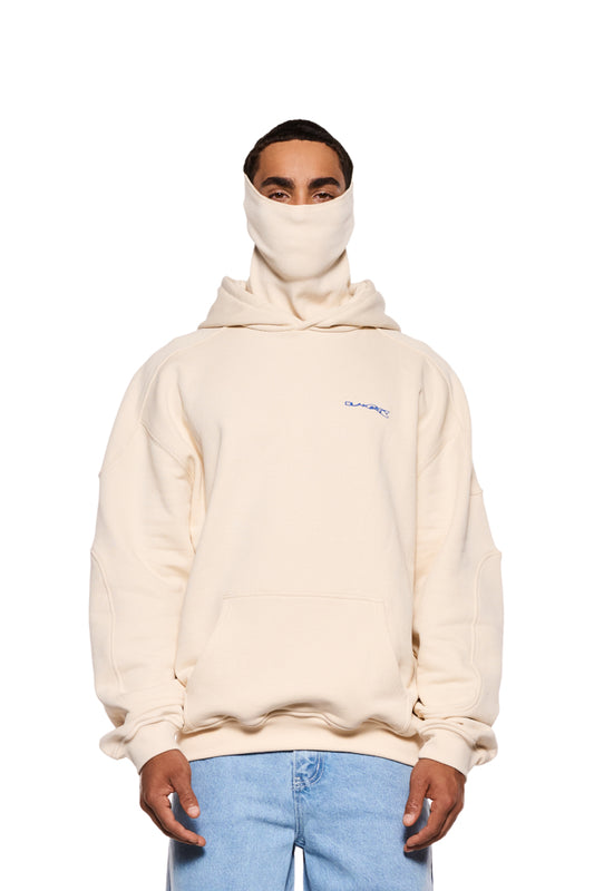 Mann trägt Synergy Mask Hoodie Beige mit abnehmbarer Maske, Teilungsnähten und Synergy-Grafik auf dem Rücken. Kombiniert mit blauen Jeans.
