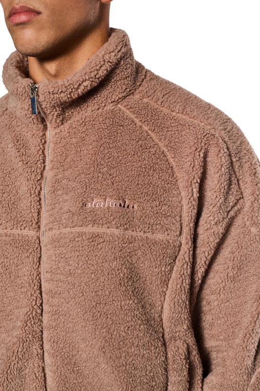 Nahaufnahme einer Person, die eine Teddy Jacket Brown mit hohem Kragen und gesticktem OLAKALA-Logo auf der Brust trägt, zeigt Teilungsnähte und elastische Bündchen.