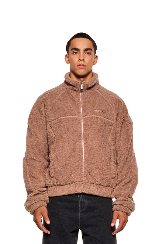 Mann trägt Teddy Jacket Brown mit hohem Kragen, Teilungsnähten und gesticktem OLAKALA-Logo. Sichtbare Eingriffstaschen und Innentasche mit Reißverschluss.