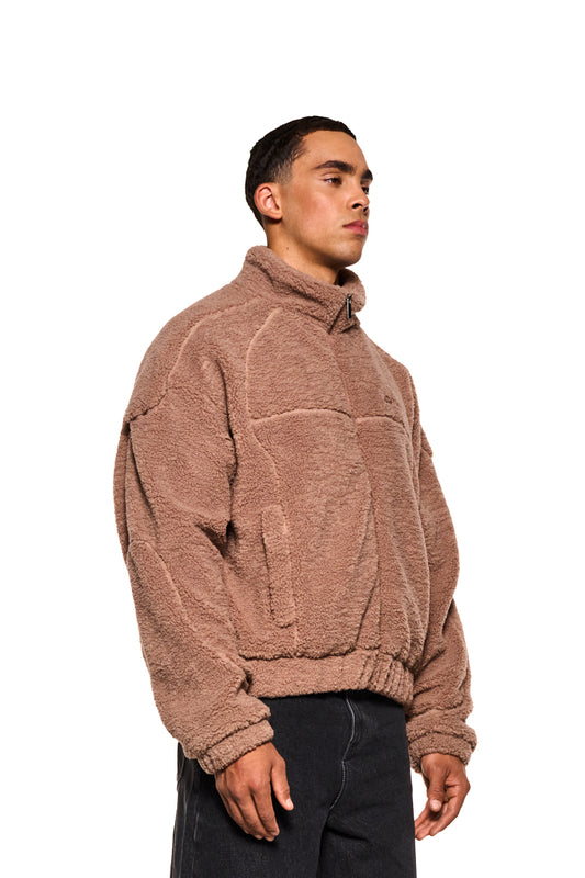 Mann in brauner Teddy Jacket mit hohem Kragen, Teilungsnähten, Eingriffstaschen, elastischen Bündchen und gesticktem OLAKALA-Logo auf der Brust.
