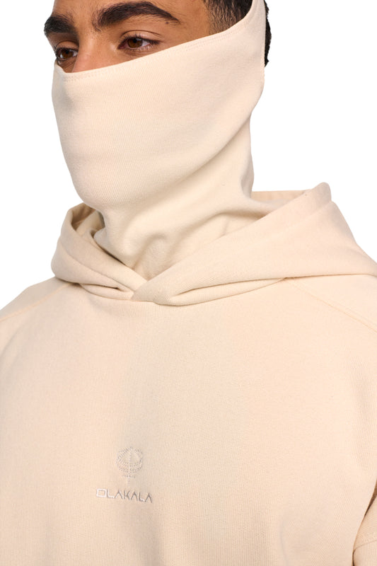 Person trägt Discreet Mask Hoodie Beige mit gesticktem OLAKALA-Logo, Ripp-Bündchen und elastischem Saum, Teilungsnähte an Schultern, Ärmeln und Kapuze sichtbar.