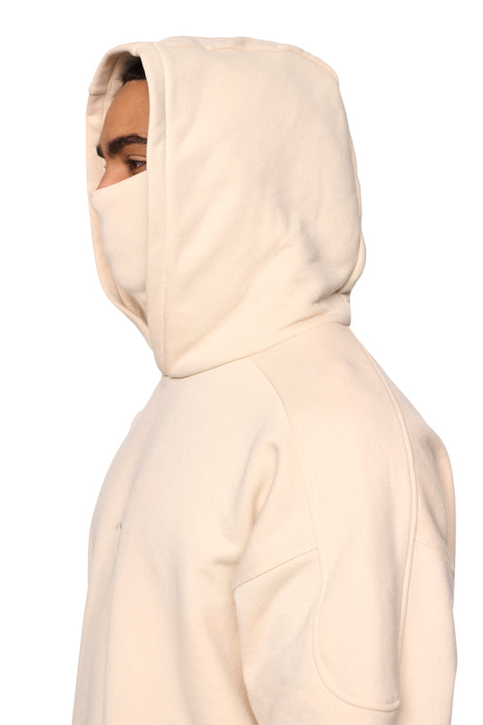 Person trägt Discreet Mask Hoodie Beige mit Kapuze, gesticktem OLAKALA-Icon auf der Brust, Ripp-Bündchen, elastischem Saum, Teilungsnähten und oversized, cropped Fit.