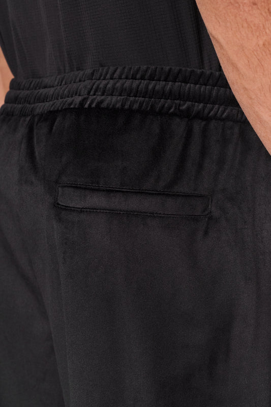 Nahaufnahme der Sports Velour Jogger Pants Black mit weichem Velours-Material, offenen Beinabschlüssen, Seitentaschen und gesticktem OLAKALA Sports Icon auf dem linken Bein.