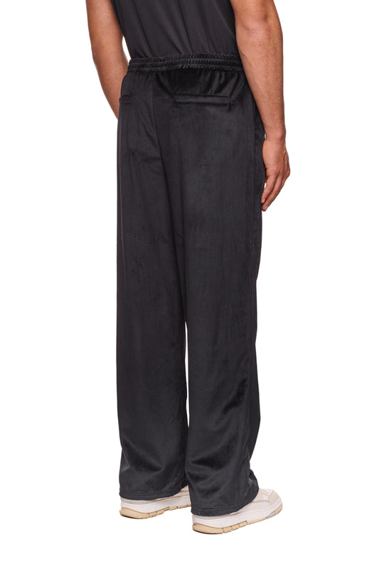 Person trägt Sports Velour Jogger Pants Black mit offener Beinabschluss, Eingriffstaschen und gesticktem OLAKALA Sports Icon auf dem linken Bein.