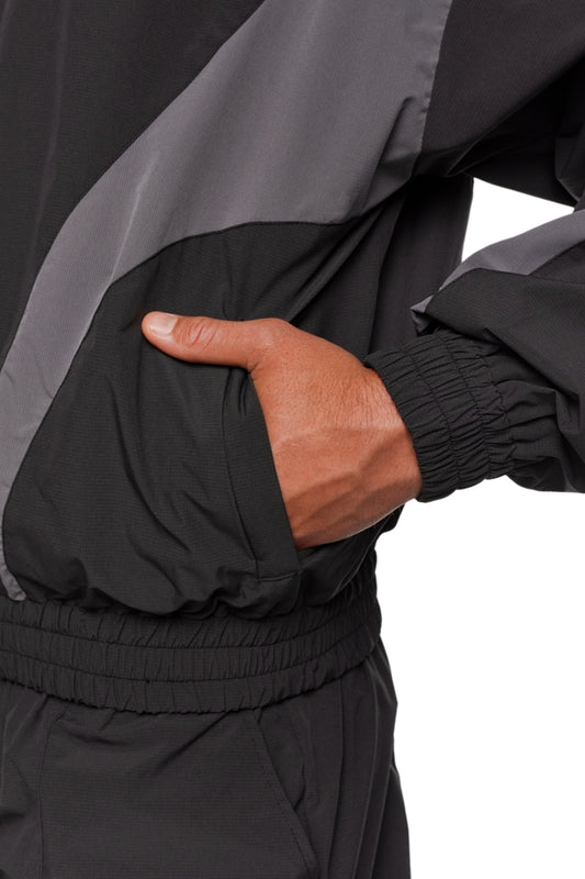 Person trägt schwarze und graue OLAKALA Sports Trackjacket mit robustem Rip-Stop Gewebe, atmungsaktivem Mesh-Futter, elastischen Bündchen und gesticktem OLAKALA Logo.