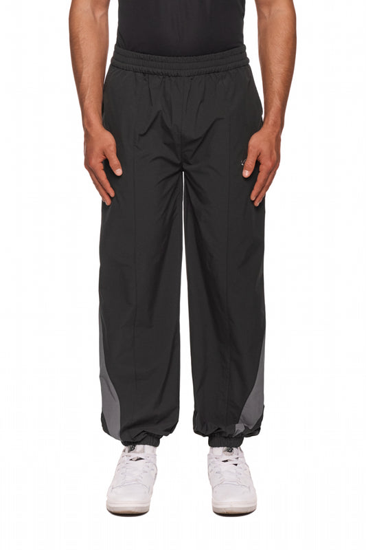 Person trägt OLAKALA Sports Trackpants Black mit elastischem Bund, kontrastfarbene Einsätze und gesticktem Logo auf linkem Bein, kombiniert mit weißen Sneakers.
