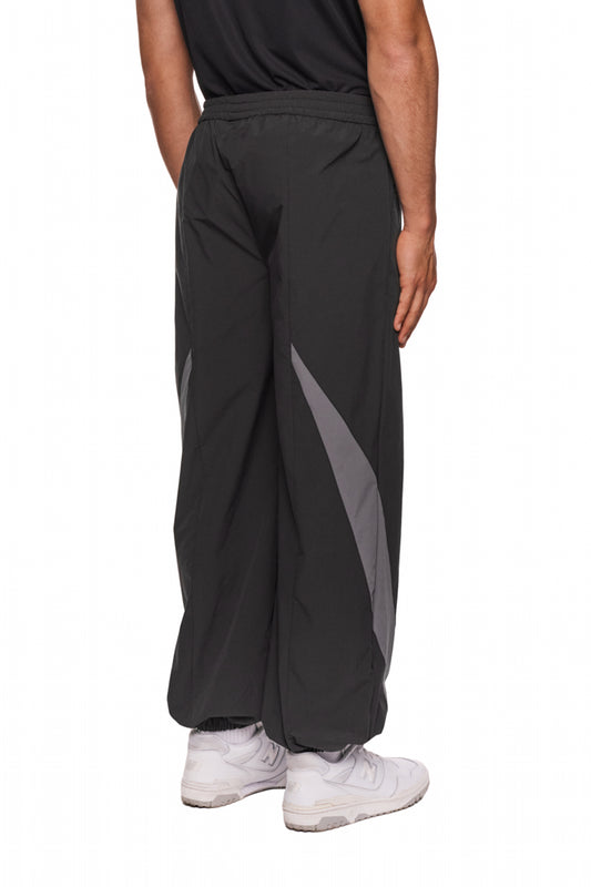 Person trägt schwarze Sports Trackpants mit gesticktem OLAKALA Sports Logo, elastischen Bündchen und kontrastierenden Stoffeinsätzen, ergänzt durch weiße Sneakers.