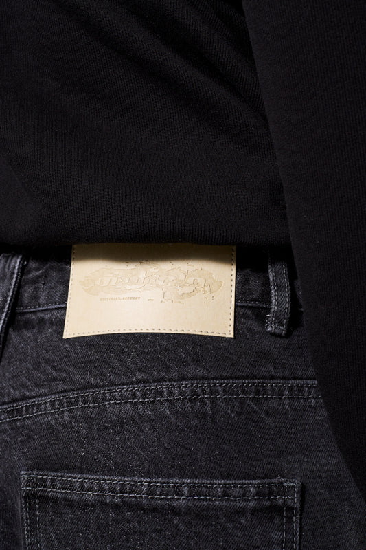 Person trägt CC Black Denim Pants Baggy mit versetztem Hosenbund und geprägtem OLAKALA-Patch, betont durch lockere Passform und einzigartigen Mittelfinger-Patch.