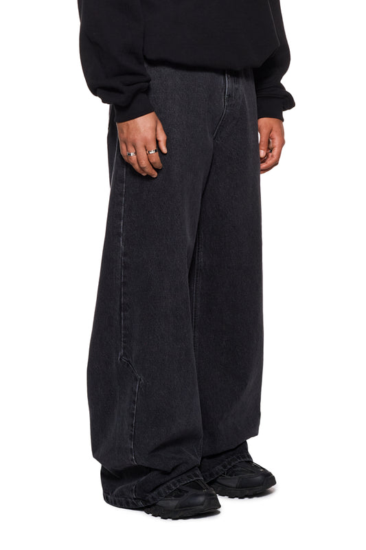 Person trägt CC Black Denim Pants Baggy von OLAKALA mit versetzten Nähten und Taschen, lockerem Schnitt, OLAKALA-Logo und charakteristischem Mittelfinger-Patch.