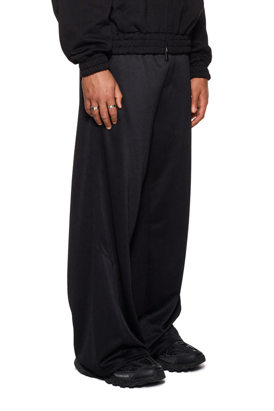 Person trägt CC Jogger Pants Black mit elastischem Bund, französischen Eingriffstaschen und gesticktem OLAKALA Logo, unisex lockere Passform. Nahaufnahme von Hand und Schuh.