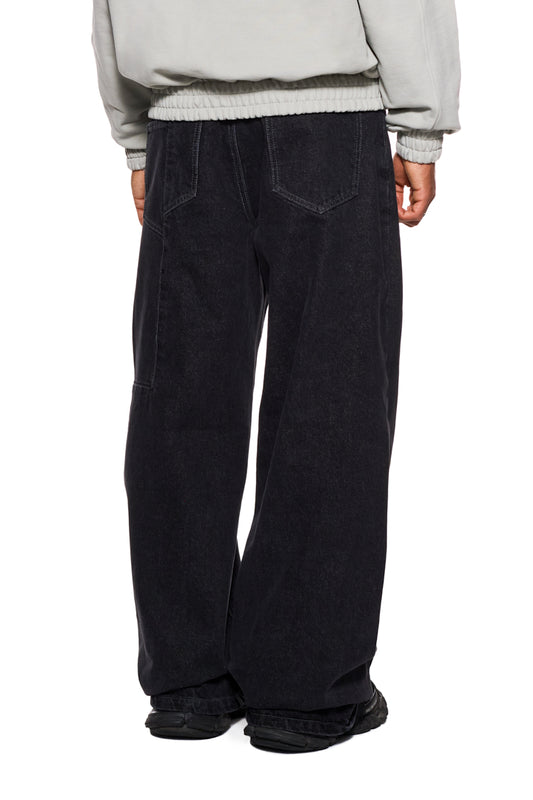 Person trägt CC Black Loose Fit Denim Pants mit versetztem Hosenbund, 5 Taschen, gesticktem OLAKALA Logo und geprägtem Patch, lockere Passform.