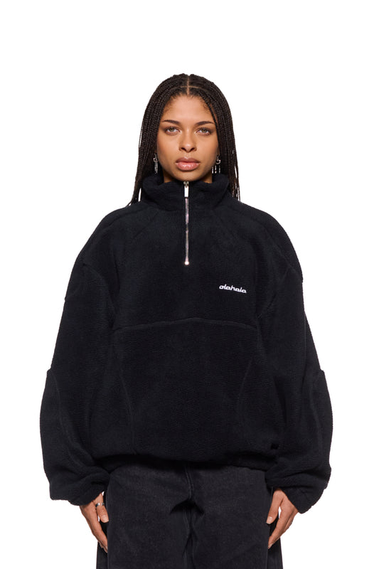 Frau trägt CC Fleece Halfzip Black mit hohem Kragen und gesticktem OLAKALA Logo, sichtbar sind Reißverschluss und elastische Bündchen.