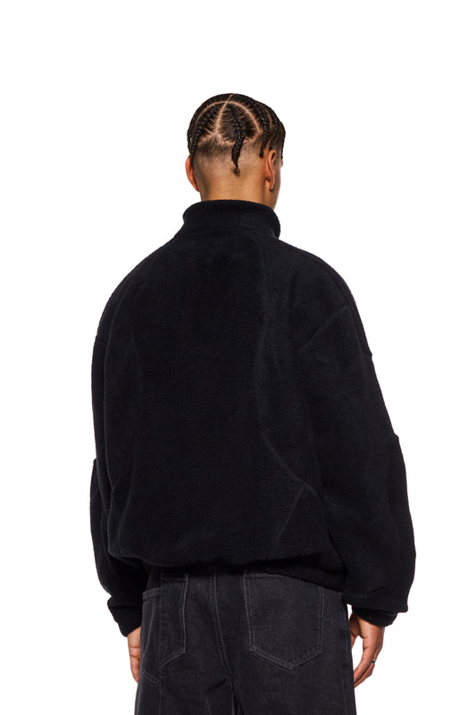 Person mit geflochtenem Haar trägt den CC Fleece Halfzip Black mit hohem Kragen und gesticktem OLAKALA Logo auf der Brust, zeigt elastische Bündchen und Reißverschluss.