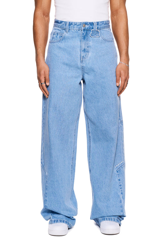 Person wears CC Blue Loose Fit Denim Pants with offset side seam, OLAKALA logo and patch. Features 5-pocket style and loose fit.