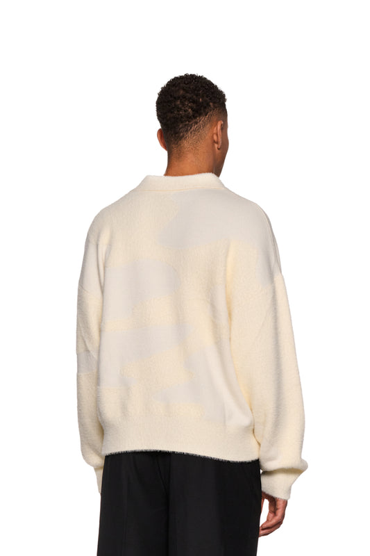 Mann trägt den Glitched Fuzzy Polo Cream mit Polo-Kragen und Synergy-Logo, kombiniert mit schwarzer Hose.