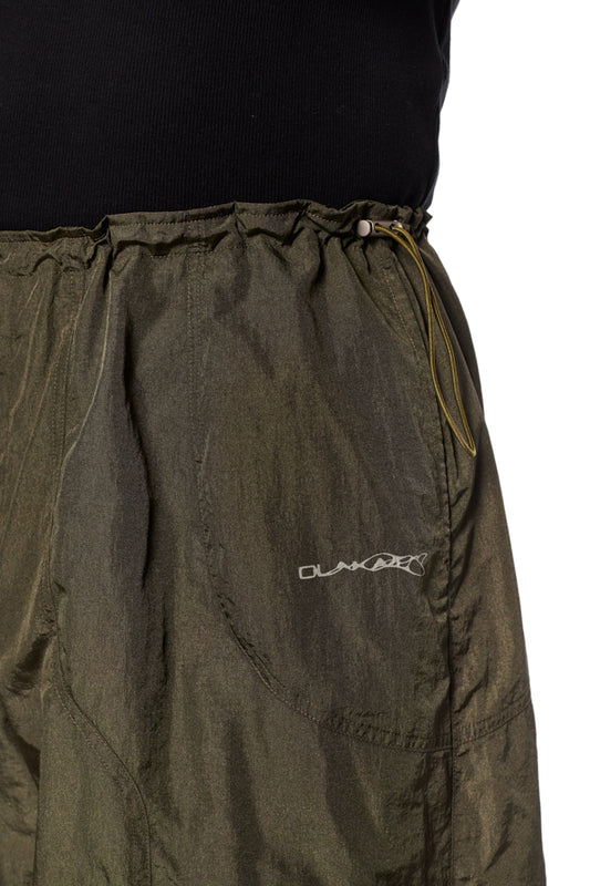 Person trägt Khakifarbene Parachute Pants mit elastischem Kordelzug, Eingriffstaschen und gesticktem Synergy-Logo, bekannt für lockeren Sitz und strapazierfähiges Material.