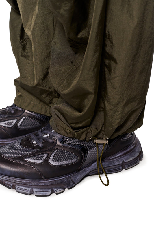 Eine Person trägt die Parachute Pants Khaki mit elastischem Kordelzug und gesticktem Synergy-Logo, kombiniert mit schwarzen Schuhen.