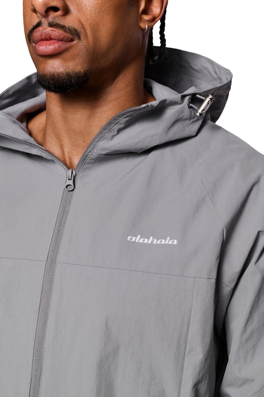 Man wears Windbreaker Light Gray with zipper, elastic cuffs, adjustable hood and embroidered OLAKALA logo on the chest.