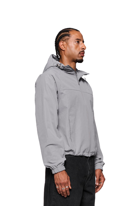 A man wears a Light Gray windbreaker with zipped pockets and embroidered OLAKALA logo, hood and elasticated sleeves.