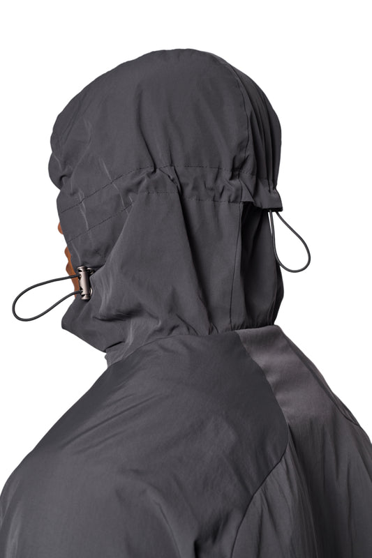 Person trägt Windbreaker Dunkelgrau mit Reißverschluss vorne, verstellbarer Kapuze, elastischen Bündchen und gesticktem OLAKALA Logo auf der Brust.