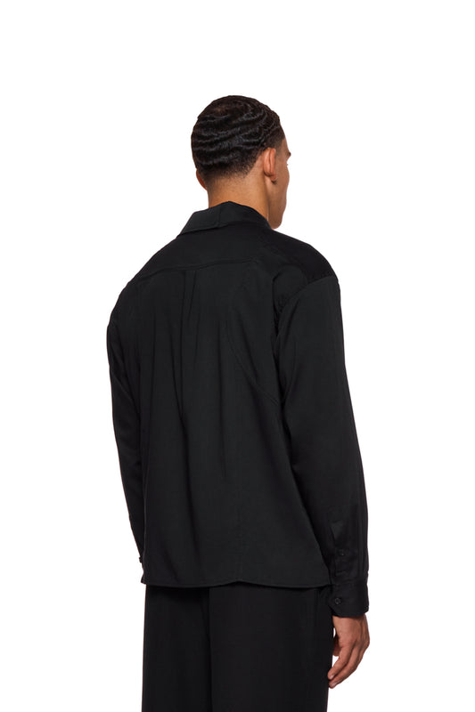 Man wears the CC Anatomy Shirt Black by OLAKALA, with button placket, double-layer collar, patch breast pocket and embroidered logo, loose fit.