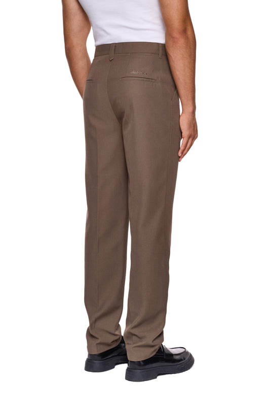 Mann trägt OLAKALA CC Dress Pants Slim Fit in Braun mit asymmetrischer Tasche und gesticktem Logo, schmale Passform, aus robustem Köper-Gewebe.