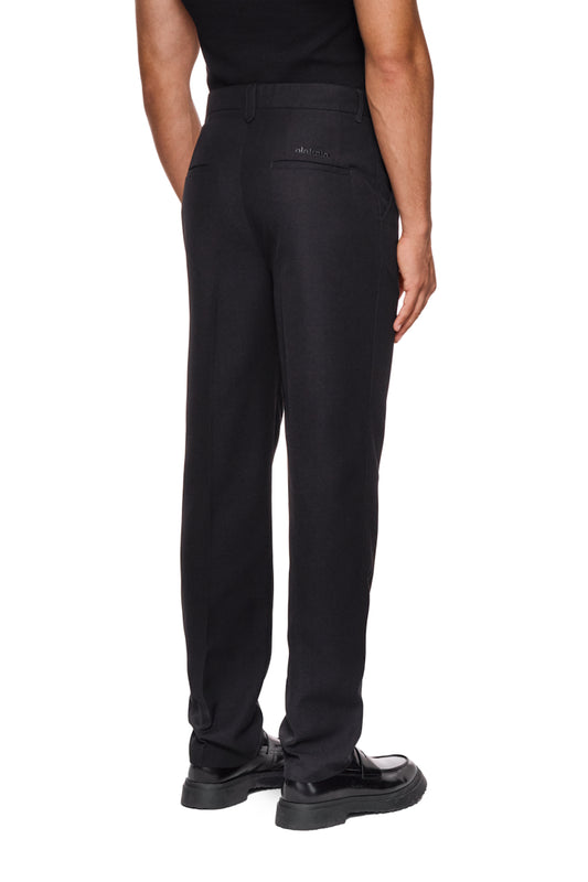 Mann trägt CC Dress Pants Slim Fit Black mit asymmetrischer Tasche und gesticktem OLAKALA Logo, schmale Passform, weiches Köper-Gewebe.