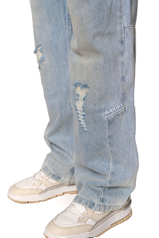 Distressed Denim Pants mit zerrissenen Stellen und hinterlegten Patches. Sichtbare gestickte OLAKALA-Logos und geprägtes Patch am Hosenbund. Sneaker im Bildausschnitt.