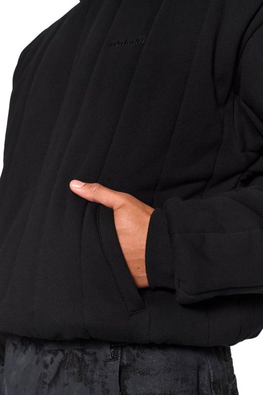 Person trägt den Greyzone Puffer Hoodie mit gesticktem OLAKALA Logo, gepufftem Design und elastischen Bündchen, während die Hand in die Jackentasche greift.