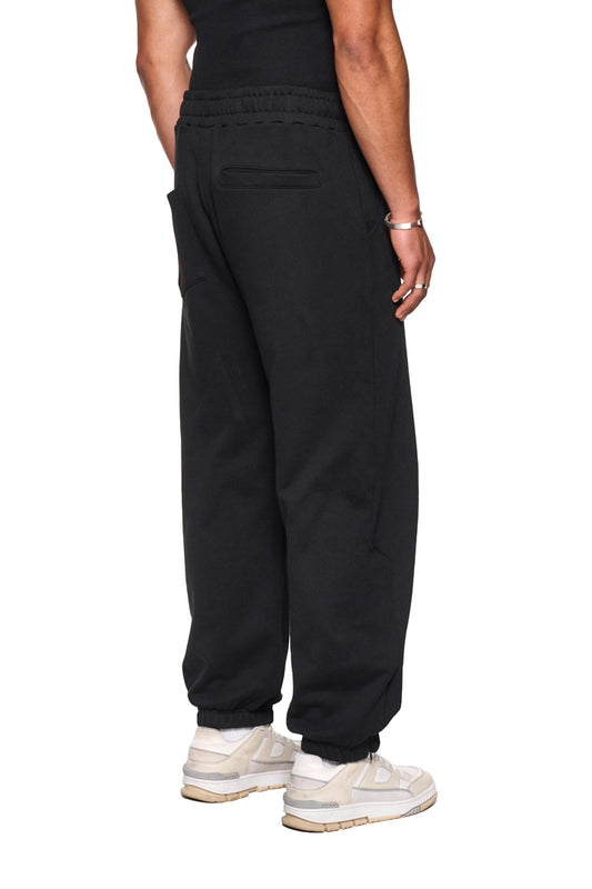 Person trägt CI Loose Sweatpants Schwarz mit elastischem Bund, asymmetrischen Taschen und gesticktem OLAKALA Logo. Lockere Passform, unisex Design, nahtloser Reißverschluss.