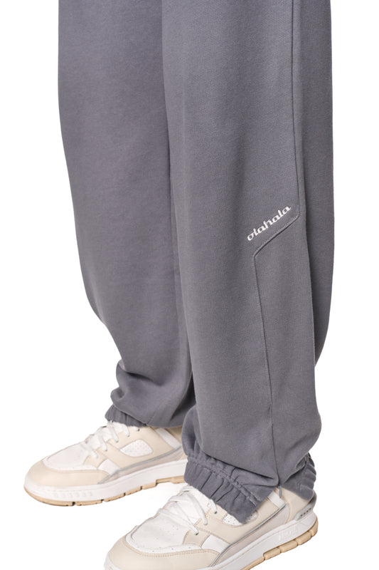 Person trägt CI Loose Sweatpants Gray mit asymmetrischen Taschen und gesticktem OLAKALA-Logo, kombiniert mit weißen Sneakers.
