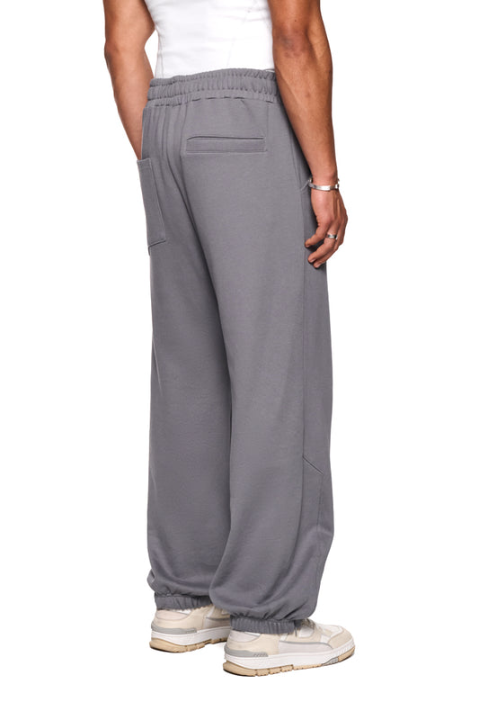 Person trägt CI Loose Sweatpants Gray mit asymmetrischen französischen Taschen und gesticktem OLAKALA-Logo, locker sitzende Jogginghose mit elastischem Bund und Kordelzug.