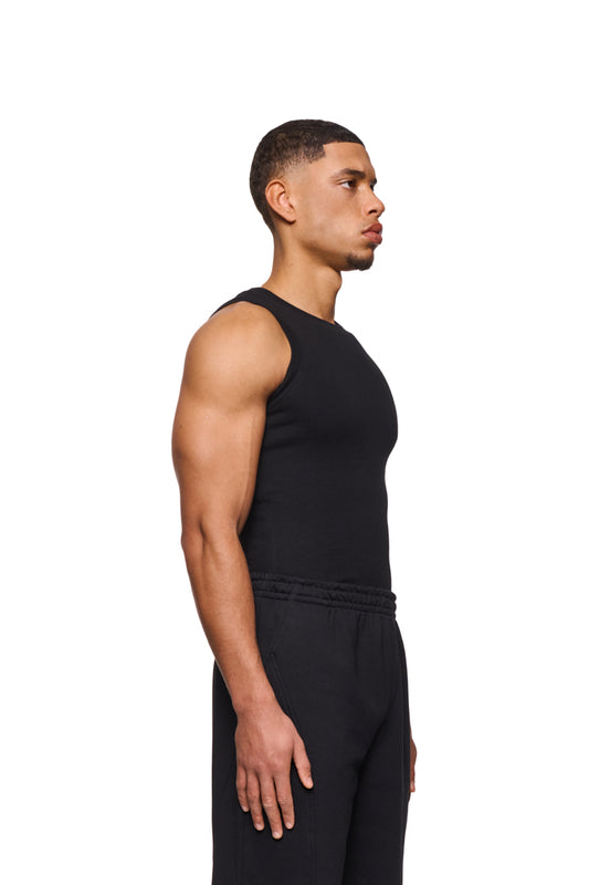 Mann im CC Core Tanktop Black, schwarzes ärmelloses Shirt mit gesticktem OLAKALA Icon auf der Brust, betont die muskulöse Passform.