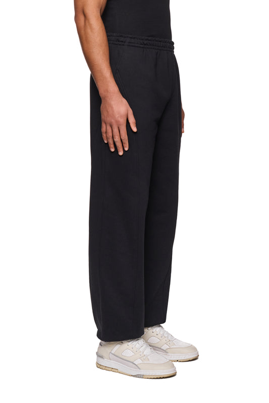 Person trägt die CC Core Sweatpants Black mit elastischem Bund und Kordelzug, kombiniert mit weißen Sneakers, Hände in den Taschen für lässigen Look.