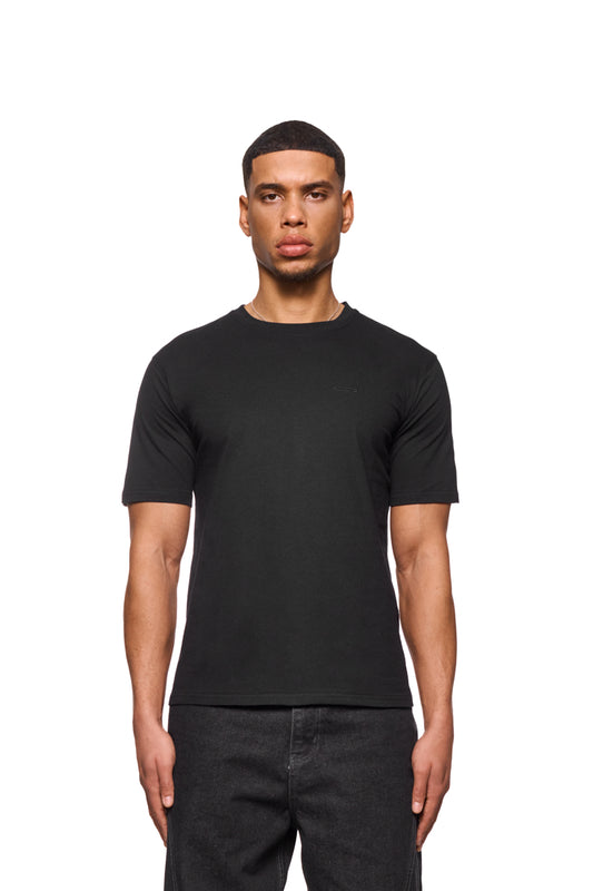Mann trägt das CC Core T-Shirt Schwarz mit Ripp-Kragen und gesticktem OLAKALA Icon auf der Brust, reguläre Passform, unisex Design.