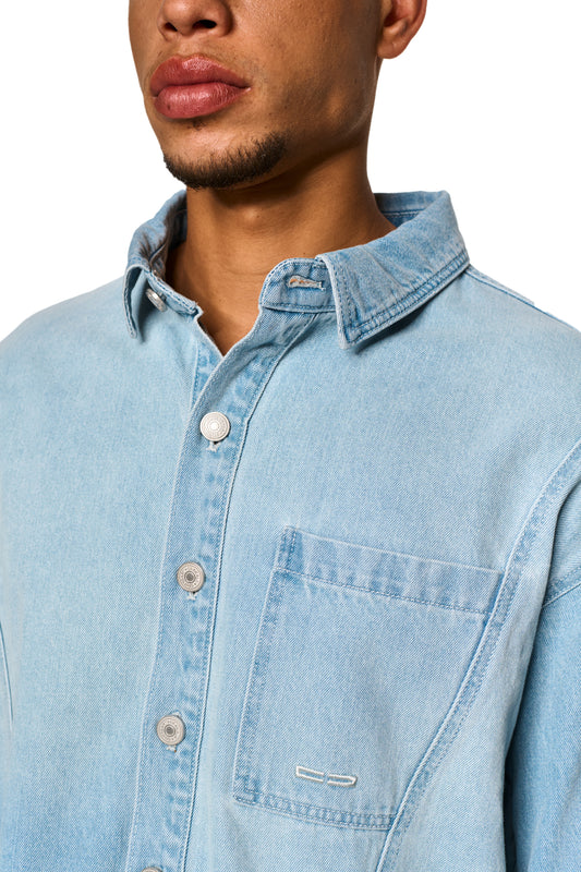 Person trägt das 0asis Denim Overshirt Blue mit Knopfleiste und OLAKALA Patch, sichtbare Teilungsnähte und Brusttaschen mit Logo-Stickerei, unisex Design.