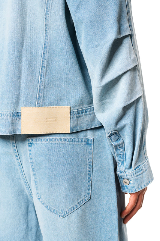 Nahaufnahme einer Hand, die die Knopfleiste eines OLAKALA Oasis Denim Overshirts Blue berührt, zeigt Details wie Brusttaschen und geprägtes Patch auf der Rückseite.