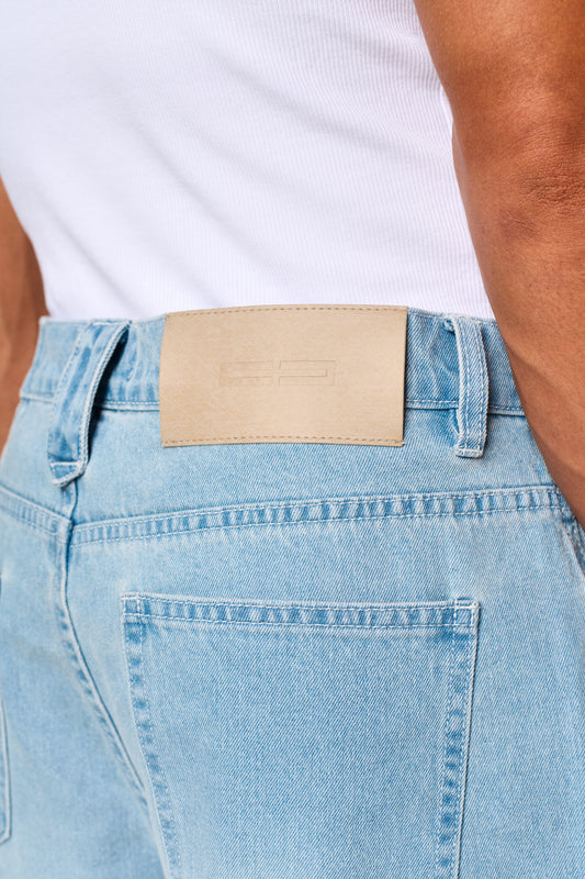 Nahaufnahme der 0asis Denim Shorts Blue mit fünf Taschen, versetzter Passe, OLAKALA Patch am Bund und Logo-Stickerei am linken Bein, reguläre Unisex-Passform.