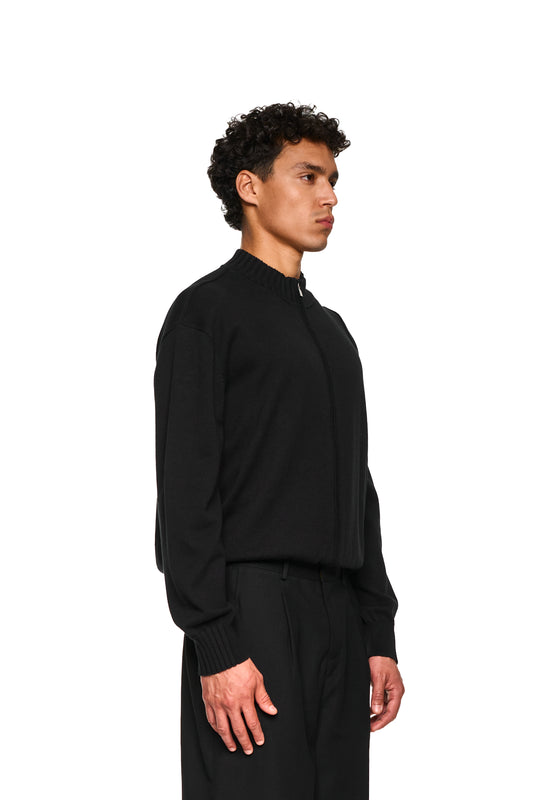 Nahaufnahme einer Hand, die den weichen Baumwollstrick der leichten Strickjacke Summer Knit Jacket Black mit asymmetrischer Vorderseite und Metallreißverschluss zeigt.