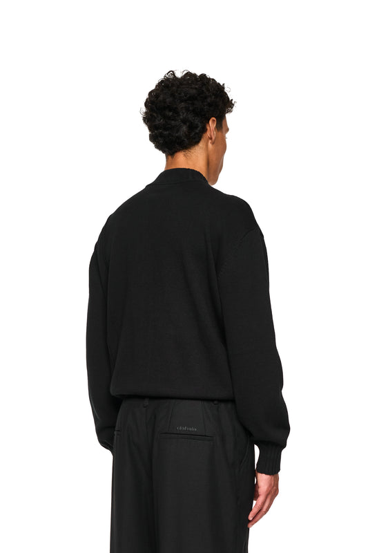 Nahaufnahme eines Mannes in einem schwarzen Pullover, der die Summer Knit Jacket Black von OLAKALA mit asymmetrischer Vorderseite und Metallreißverschluss trägt.