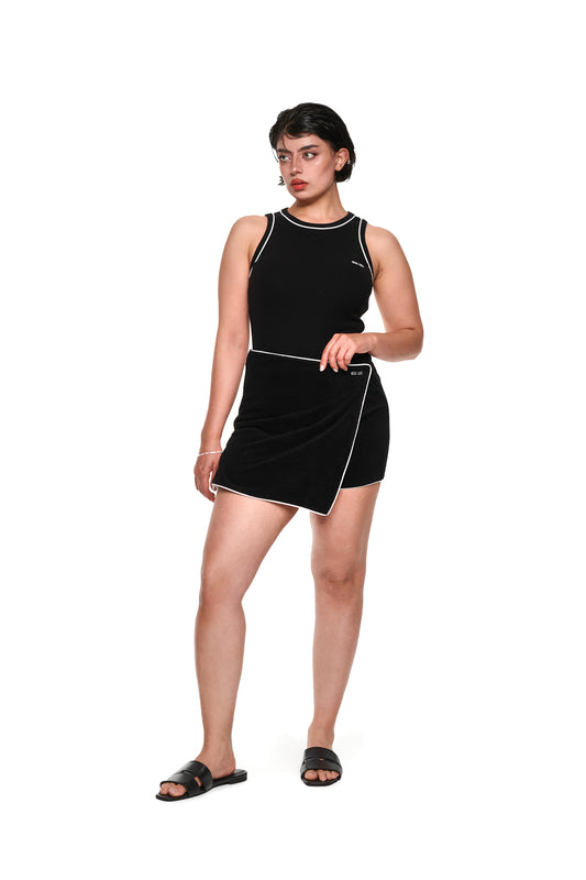 Eine Frau trägt das Contrast Tanktop Black mit elastischem Ripp-Stoff und gesticktem OLAKALA-Icon auf der Brust, betont durch Kontrast-Paspellierungen.