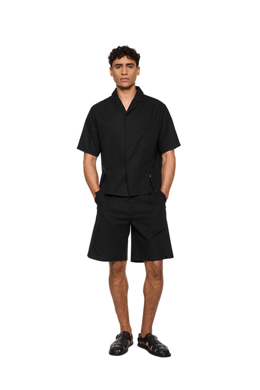 Schwarze Leinen-Shorts mit Bundfalten, französischen Taschen, asymmetrischem Eingriff, rückseitigen Leistentaschen und seitlichen Schlitzen, aus leichtem, atmungsaktivem Gewebe. Unisex, reguläre Passform.