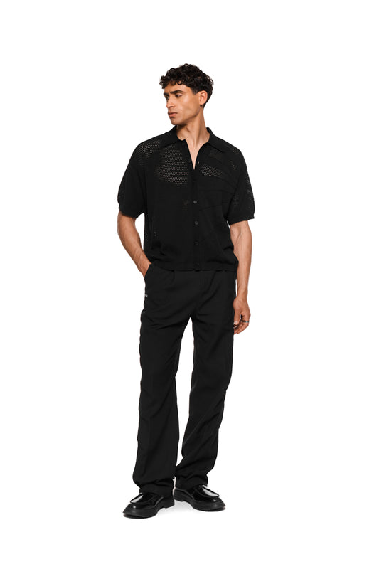 Sun Knit Shirt Black mit feinem Lochstrickmuster, Sonnendetail vorne links, doppellagigem Kragen rechts, Knopfleiste und lockerer Unisex-Passform, ideal für warme Tage.