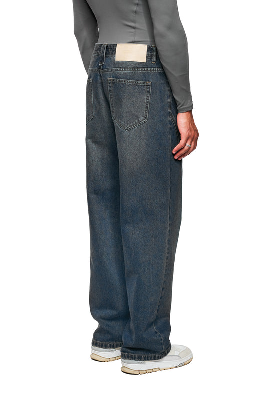 Person trägt CC Core Denim Pants Tinted Indio; weite, hochgeschnittene Jeans mit asymmetrischen Gesäßtaschen, kombiniert mit grauem Top und weißen Chunky-Sneakern.