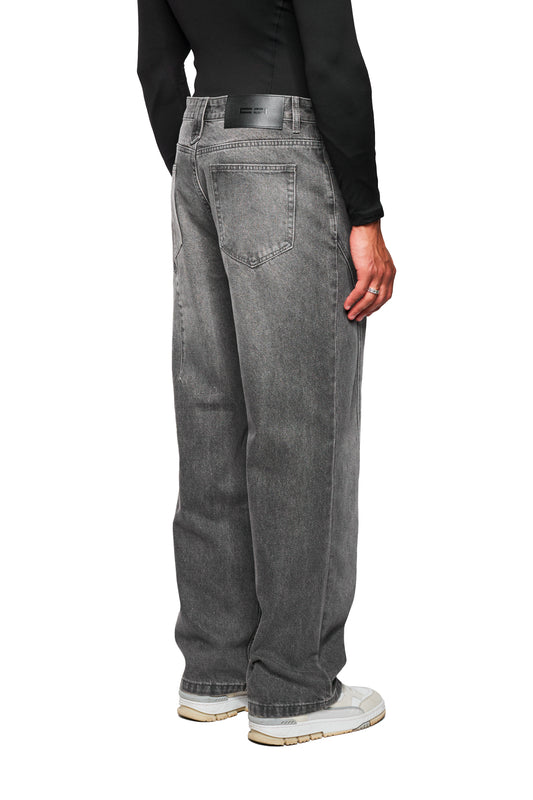 Person trägt CC Core Denim Pants Washed Gray: lockerer Schnitt, mittlere Leibhöhe, versetzte Seitennaht, asymmetrische Gesäßtaschen. Kombiniert mit cremefarbenen Sneakers.