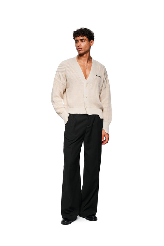 Mann trägt CC Cardigan Beige mit tiefem V-Ausschnitt, versetztem Saum, Metallpin, oversized und cropped, kombiniert mit lockerer, schwarzer Hose und schwarzen Lederschuhen.