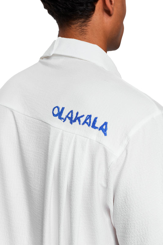 Person trägt das Crayon Summer Shirt Graphic mit blauer OLAKALA-Stickerei auf der Schulterpasse, gekennzeichnet durch kreppartige Textur und kubanischen Kragen.